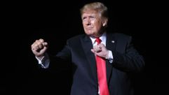 Donald J. Trump fists up ready to fight the truth at a rally in Iowa in October 2021. He is wearing a black suit jacket and red tie and is squinting into the spotlight. by the Forward
			