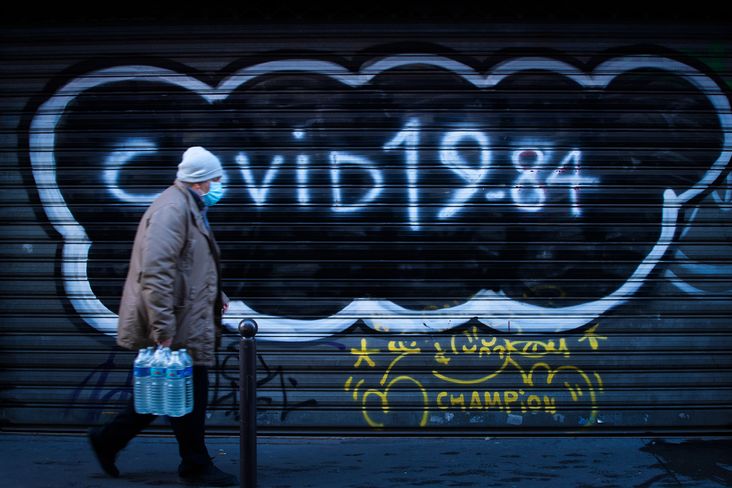 A masked pedestrian passes a gated shop storefront graffitied with the words "COVID 19-84."