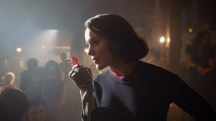 Midge Maisel in a poorly lit club, holding a cigarette and leaning into an old-timey mic.