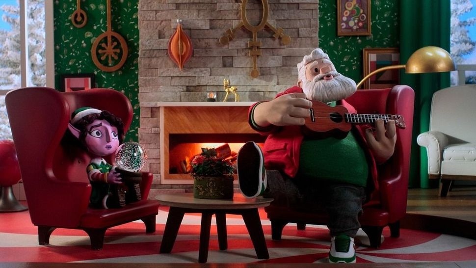 Candy the elf and Santa sit in front of a fire in red armchairs. Candy holds a snow globe and Santa holds a ukelele. She is in an elf uniform, he is in sweats and sneakers.